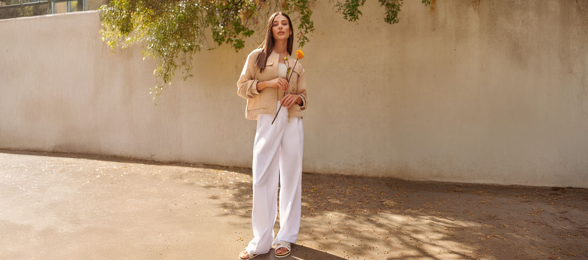 Frau in beiger Jacke, weißer Hose und Sandalen, hält eine gelbe Blume.