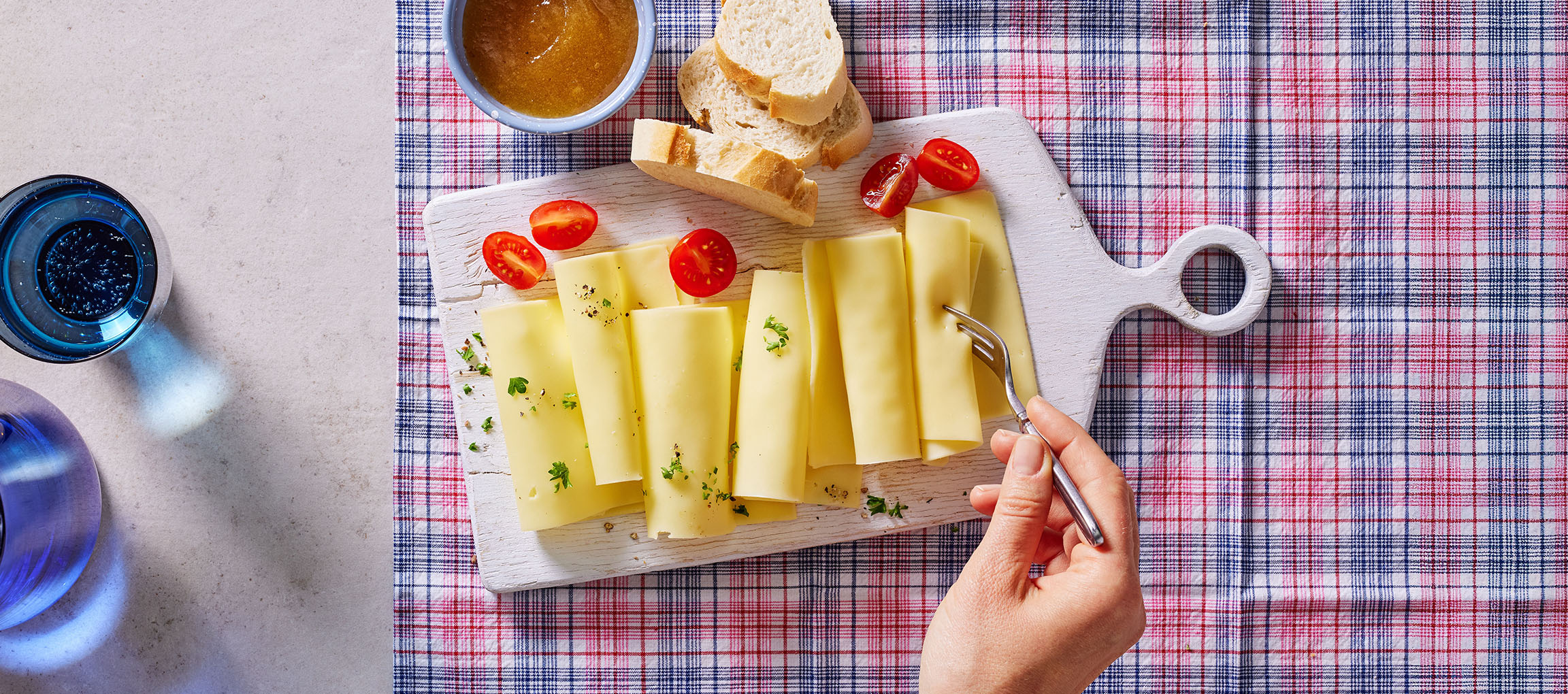 Käsescheiben mit Kirschtomaten, Brot und Marmelade auf einem karierten Tischtuch.