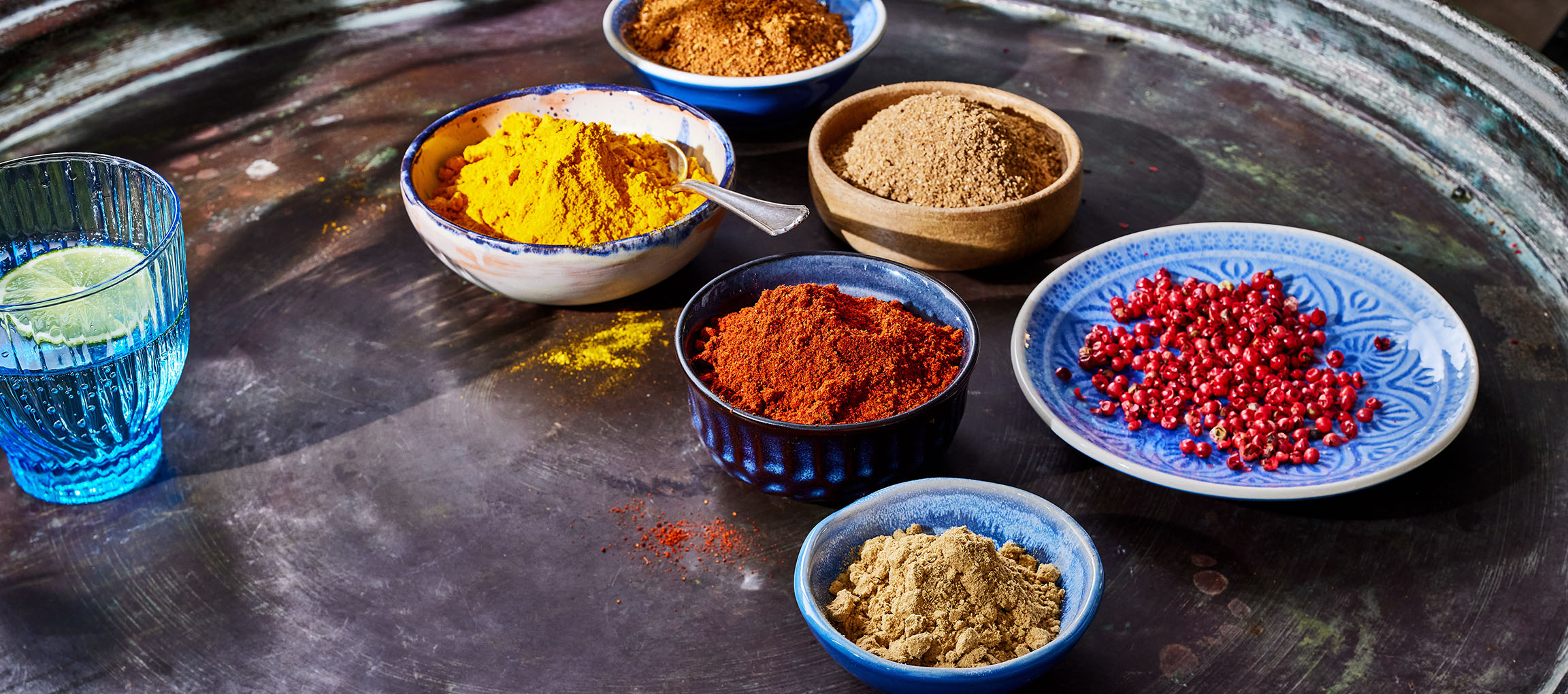 Verschiedene Gewürze, darunter Kurkuma, Paprika und rosa Pfeffer, in Schalen auf dunklem Untergrund.