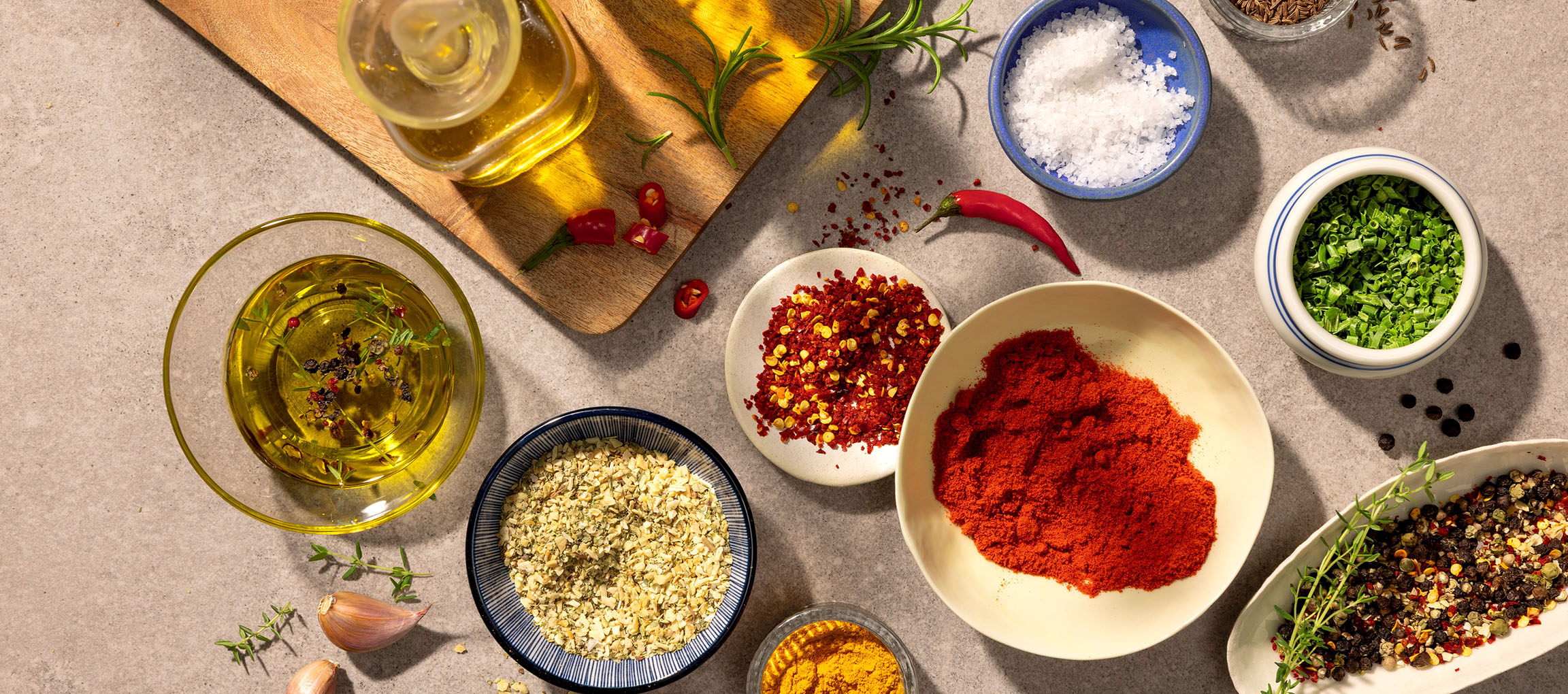 Verschiedene Gewürze und Öle, darunter Paprika, Chili, Salz, Pfeffer und Olivenöl, auf grauem Hintergrund.