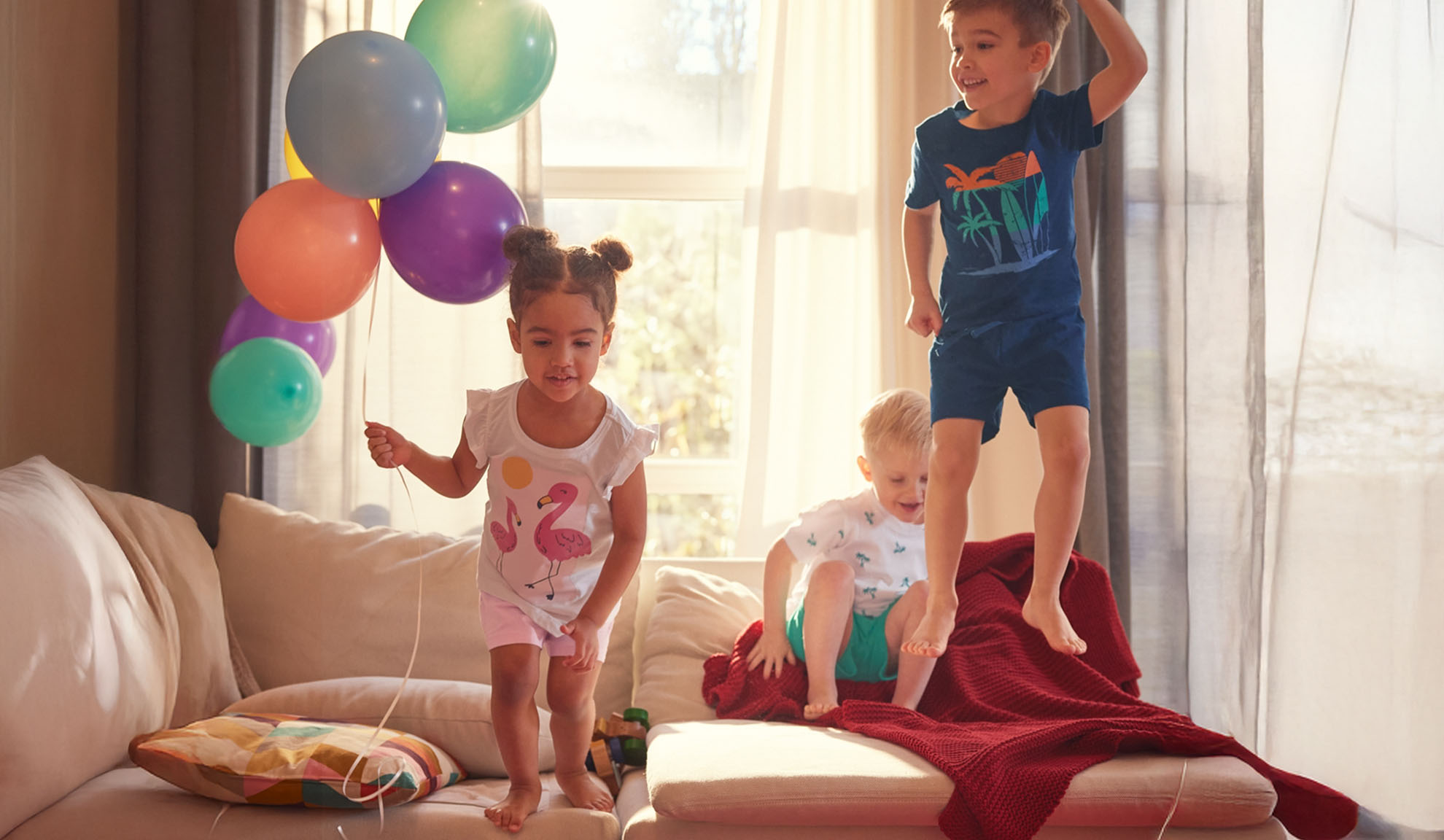Drei Kinder springen fröhlich auf einem Sofa mit bunten Ballons herum.