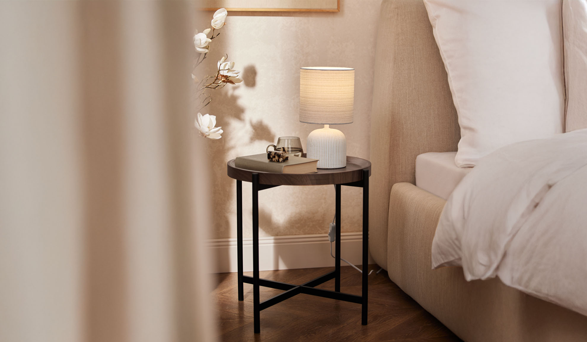 Runder Holznachttisch mit Tischlampe und Accessoires neben einem Bett.