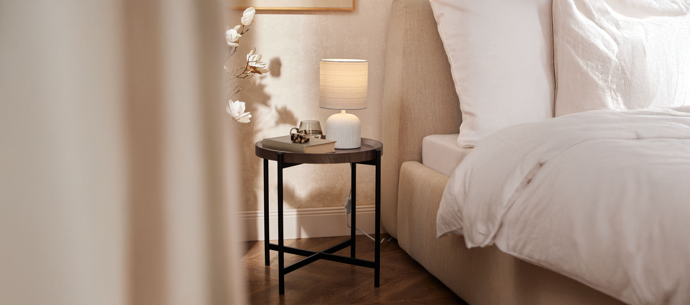 Runder Holznachttisch mit Tischlampe und beigem Bett in einem gemütlichen Schlafzimmer.