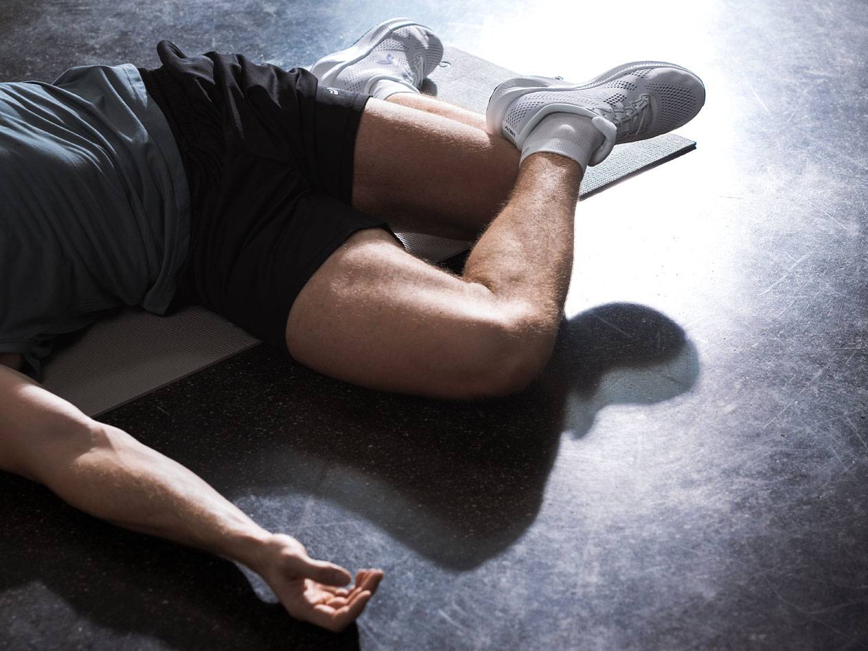 Mann liegt auf einer Gymnastikmatte in Sportkleidung und Schuhen.