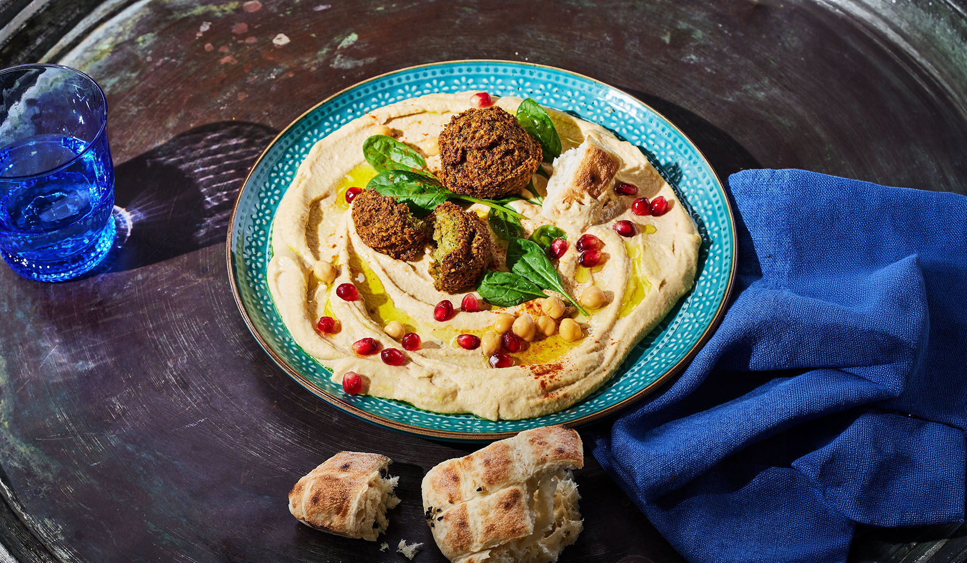 Hummus mit Falafel, Granatapfelkernen und Pitabrot auf einem dunklen Tisch.