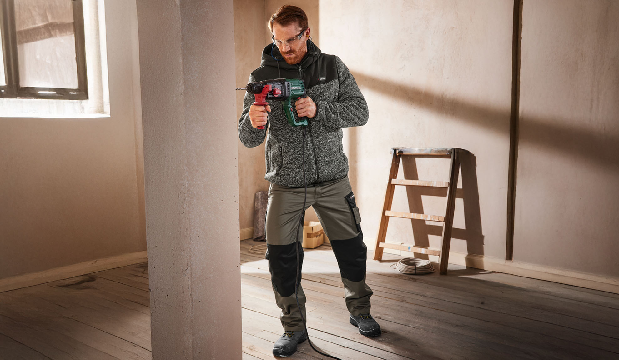Mann in Arbeitsjacke und -hose, hält einen Bohrer in einem unfertigen Raum.