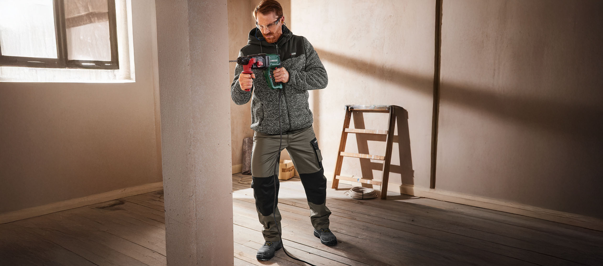 Mann in Arbeitskleidung bohrt mit Bohrmaschine in Wand.