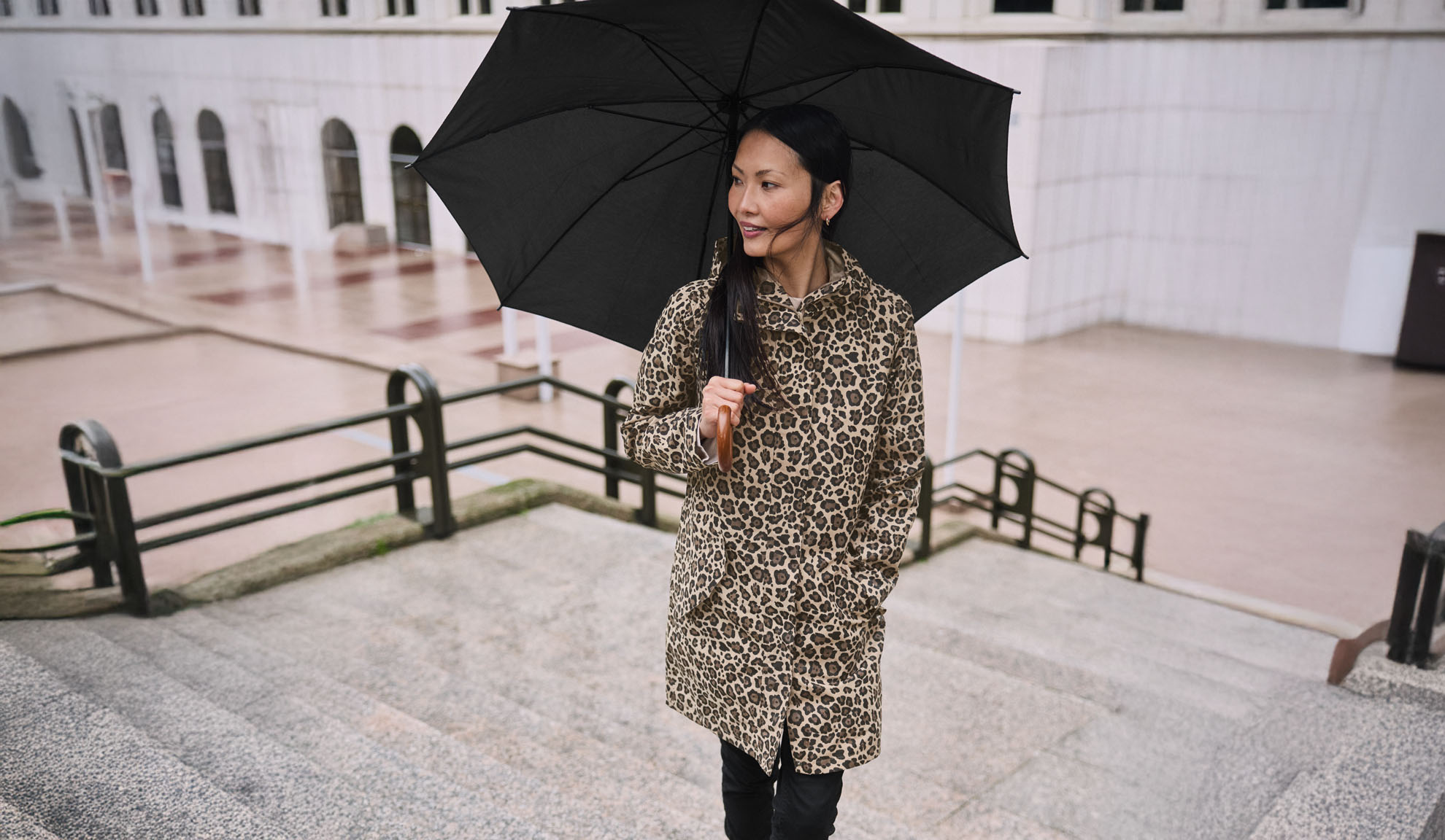 Eine Frau in einem Leopardenmantel hält einen schwarzen Regenschirm auf einer Treppe.