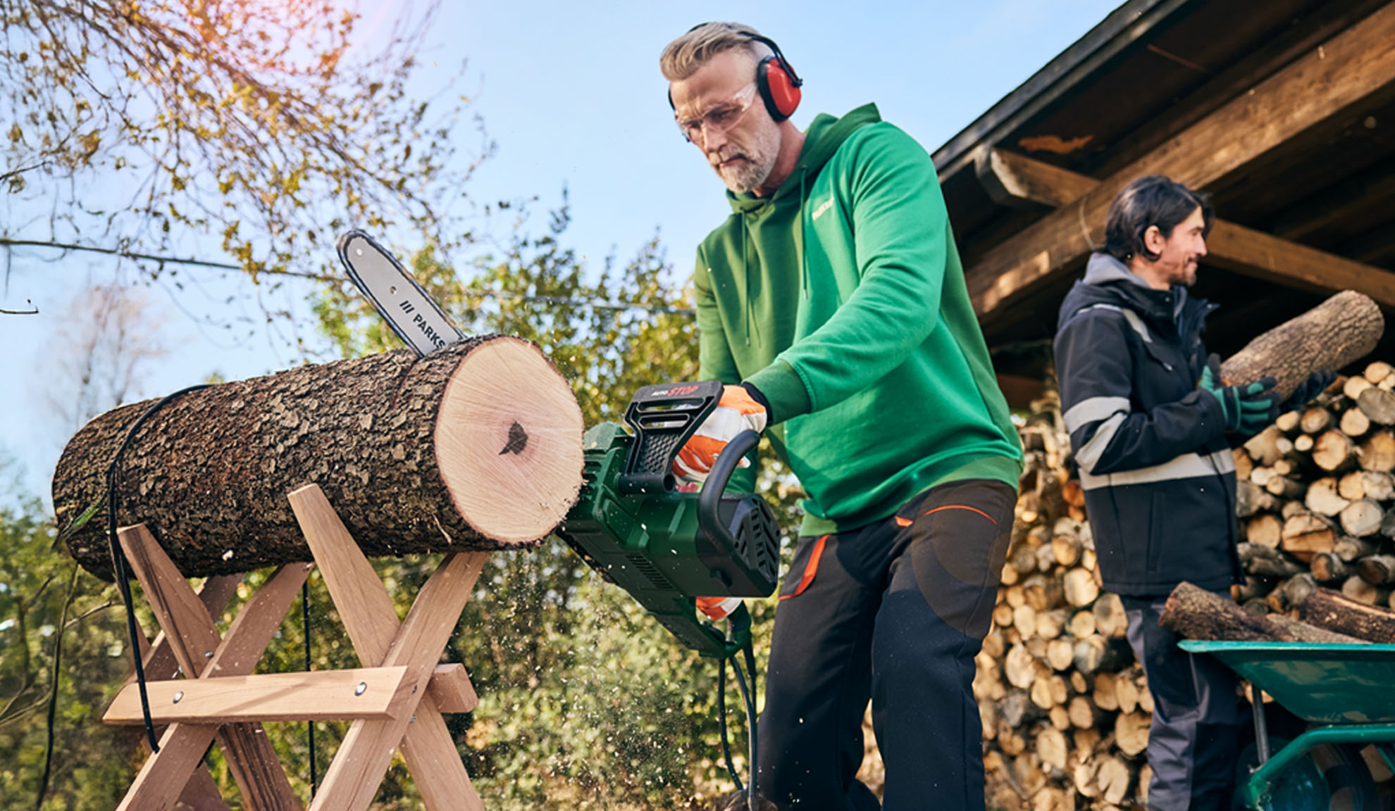 Mann sägt Baumstamm mit Kettensäge, während ein anderer Mann Holz stapelt.
