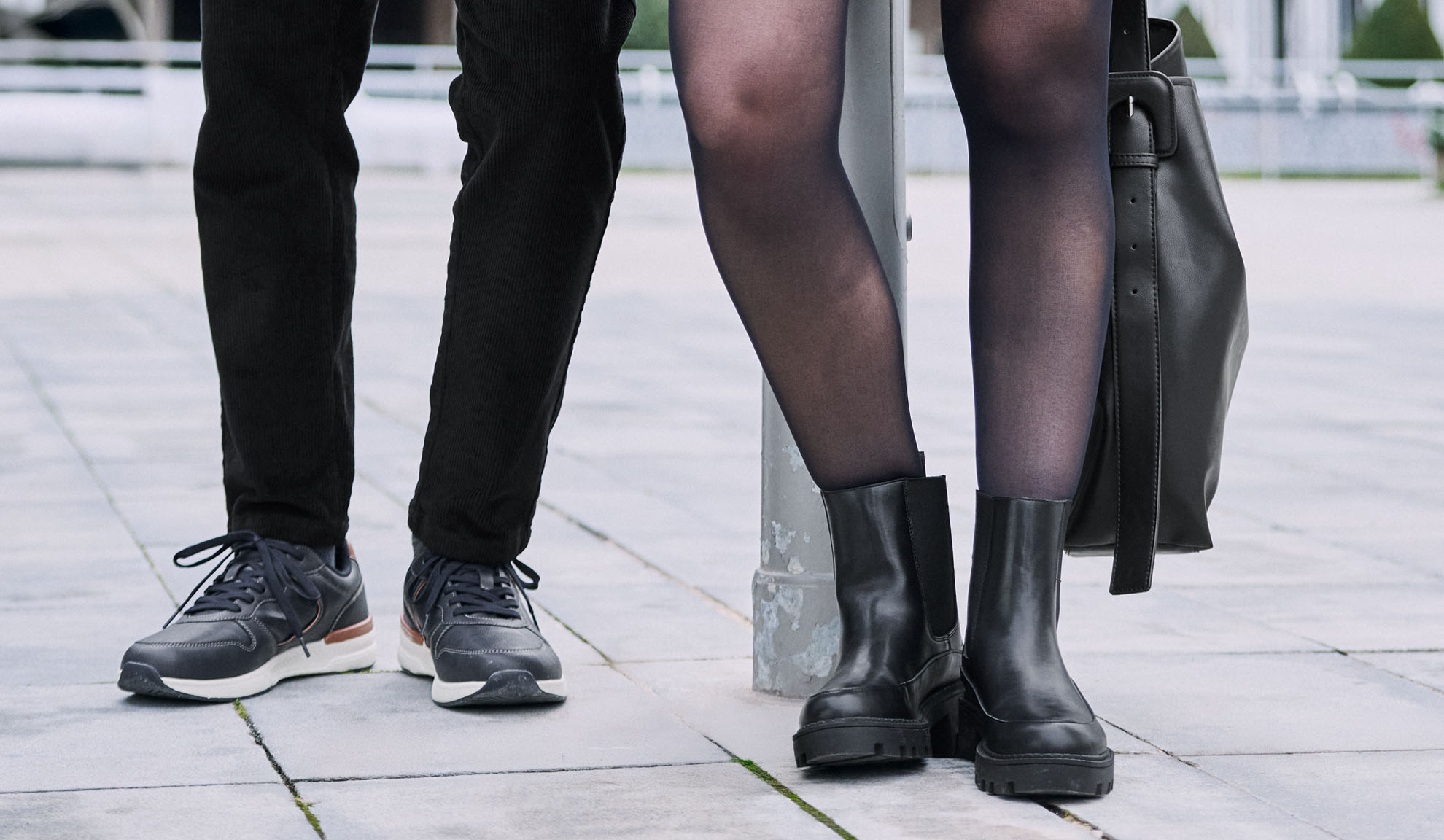 Beine von zwei Personen, eine trägt Hose und Turnschuhe, die andere Strumpfhose, Stiefel und eine Tasche.