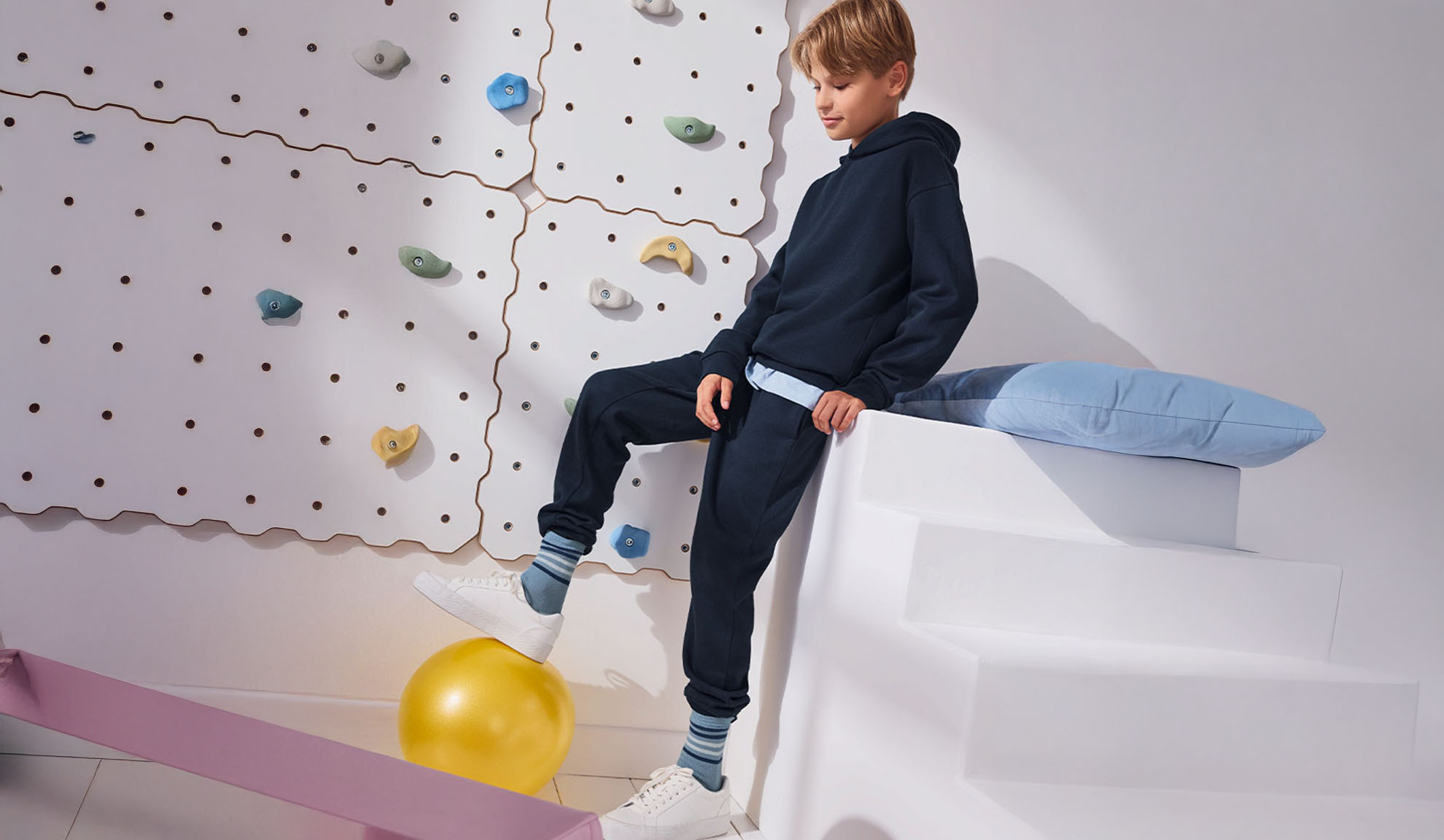 Ein Junge steht auf einem Gymnastikball neben einer Kletterwand, bekleidet mit Kapuzenpullover und Jogginghose.