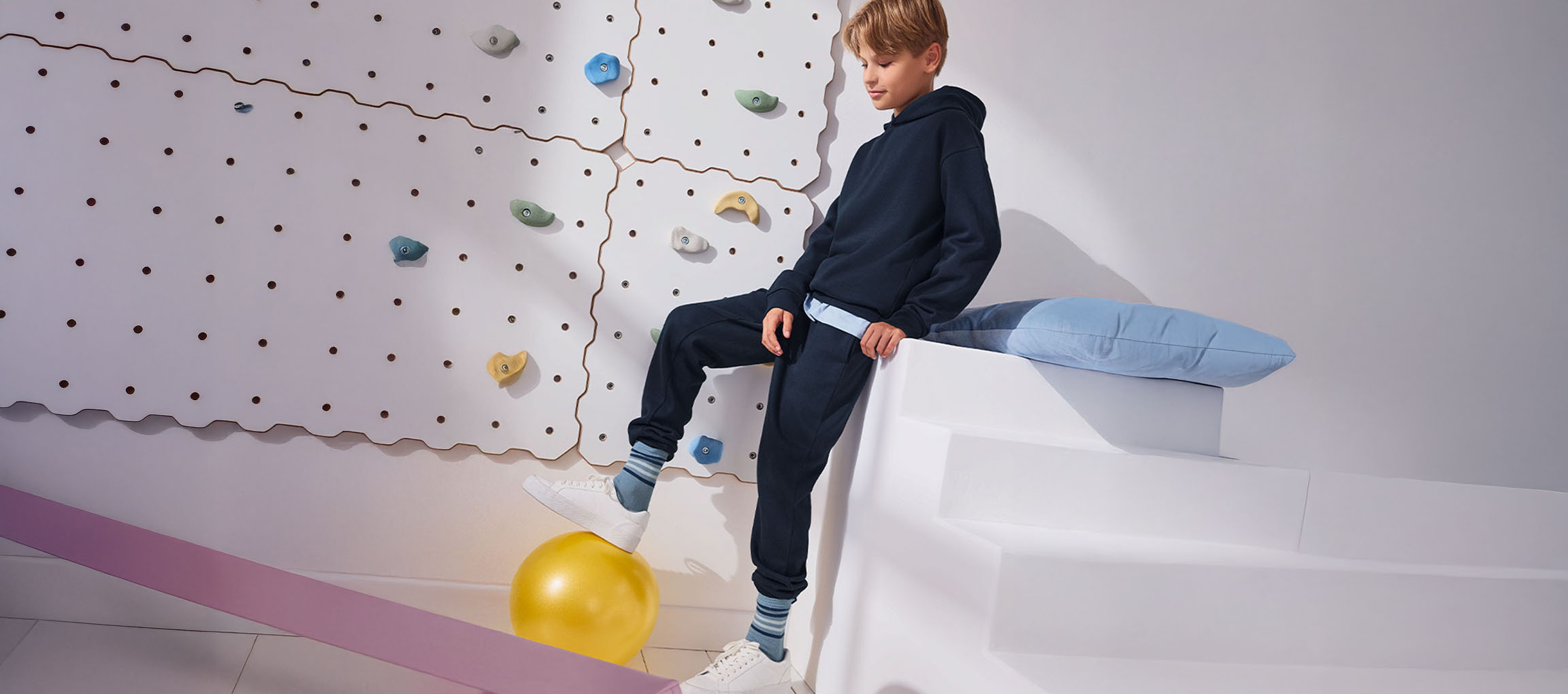 Ein Junge in marineblauem Hoodie und Jogginghose, mit einem Fuß auf einem gelben Ball, neben einer Kletterwand.