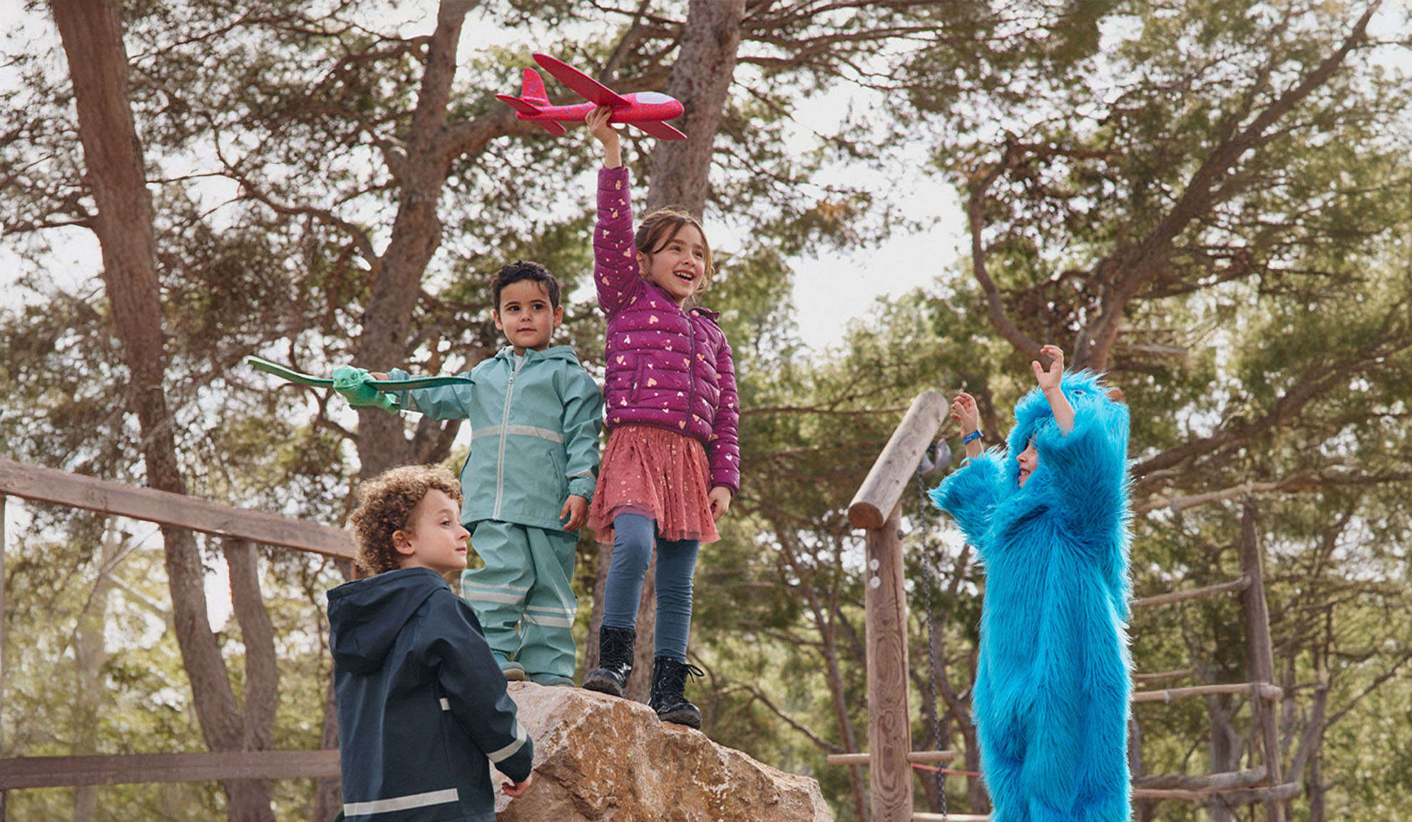 Vier Kinder spielen draußen, zwei mit Spielzeugflugzeugen und eines in einem blauen Monsterkostüm.