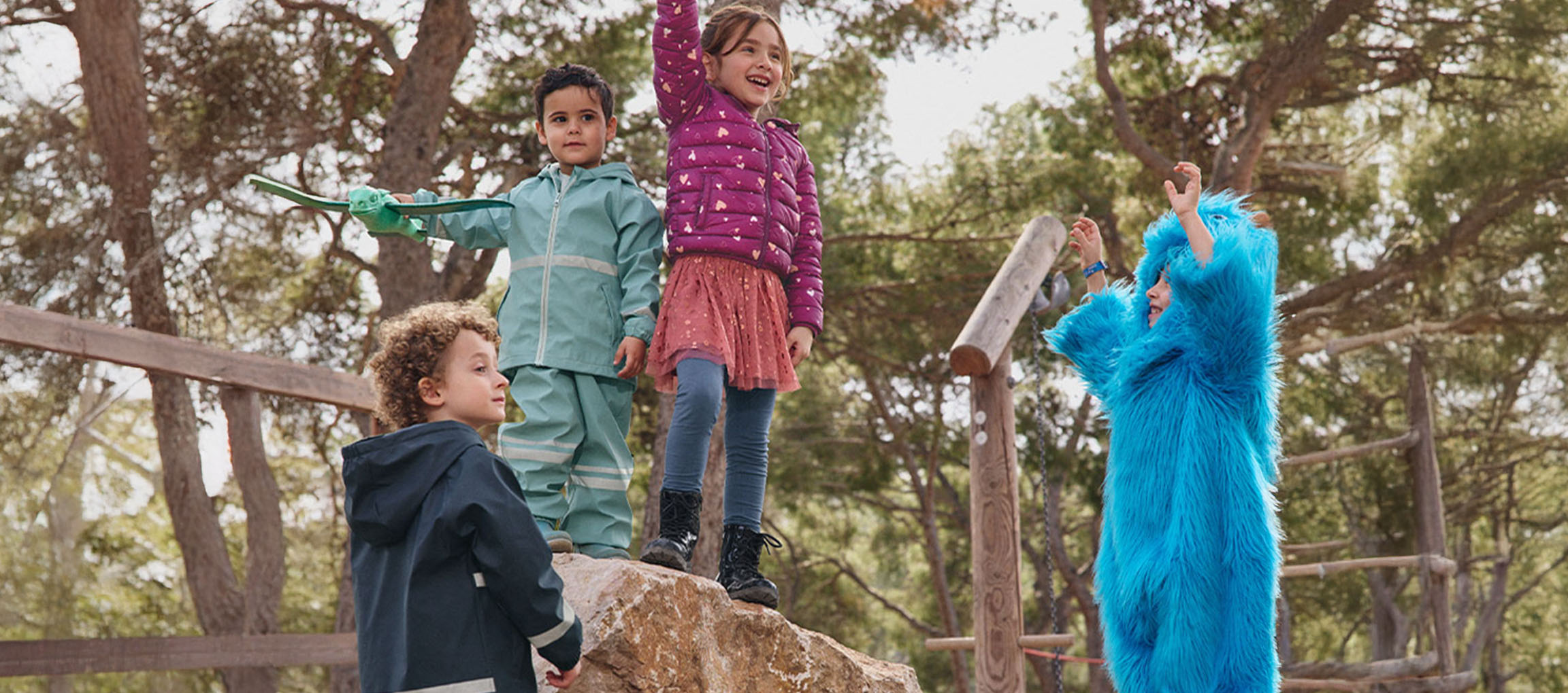 Vier Kinder spielen draußen, zwei in Oberbekleidung, eines in einem blauen Monsterkostüm.