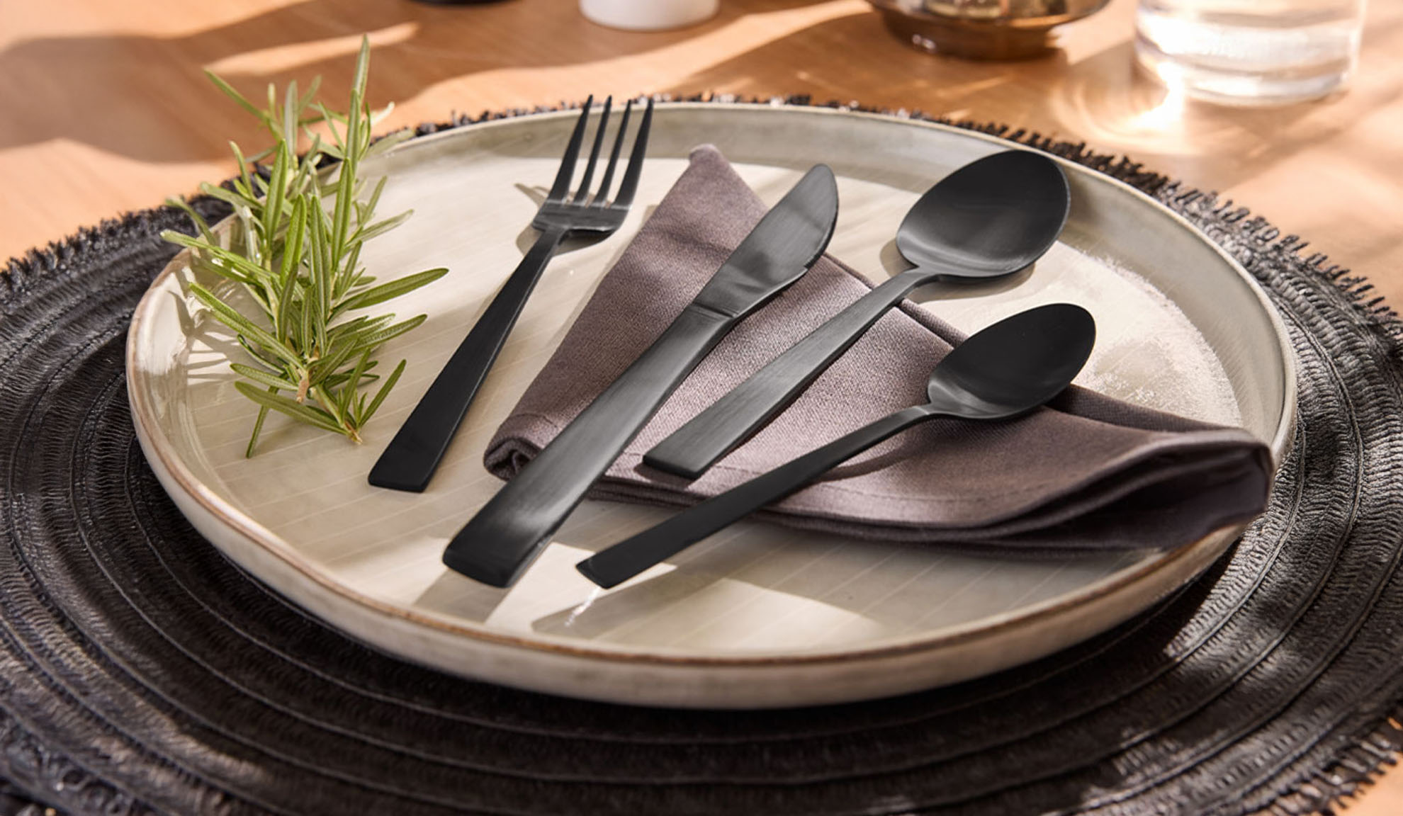 Schwarzes Besteckset mit Teller, Serviette und Rosmarinzweig auf einem Platzset.