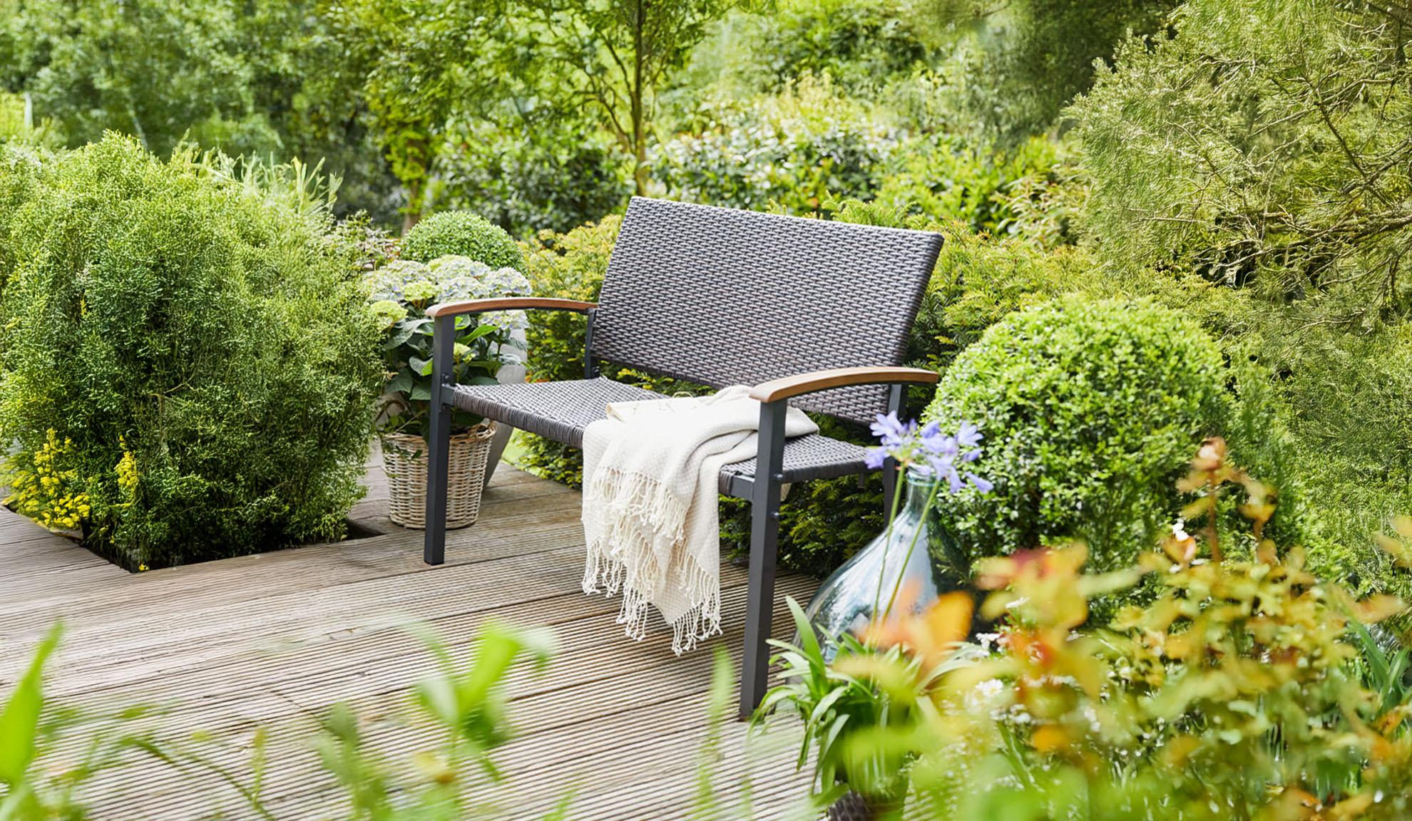 Rattan-Gartenbank auf einer Holzterrasse, umgeben von üppigem Grün.