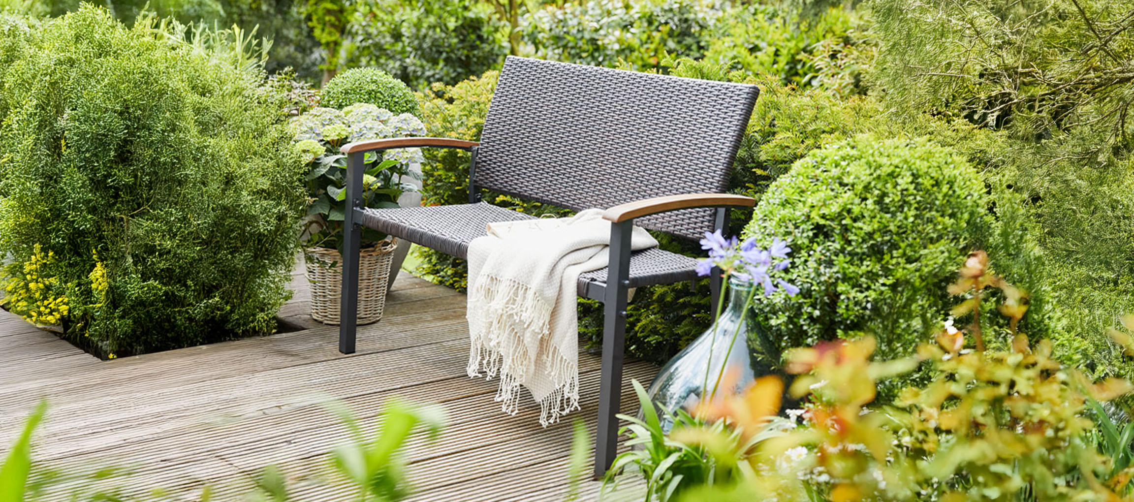 Eine graue Gartenbank mit Decke auf einer Holzterrasse, umgeben von grünen Pflanzen.
