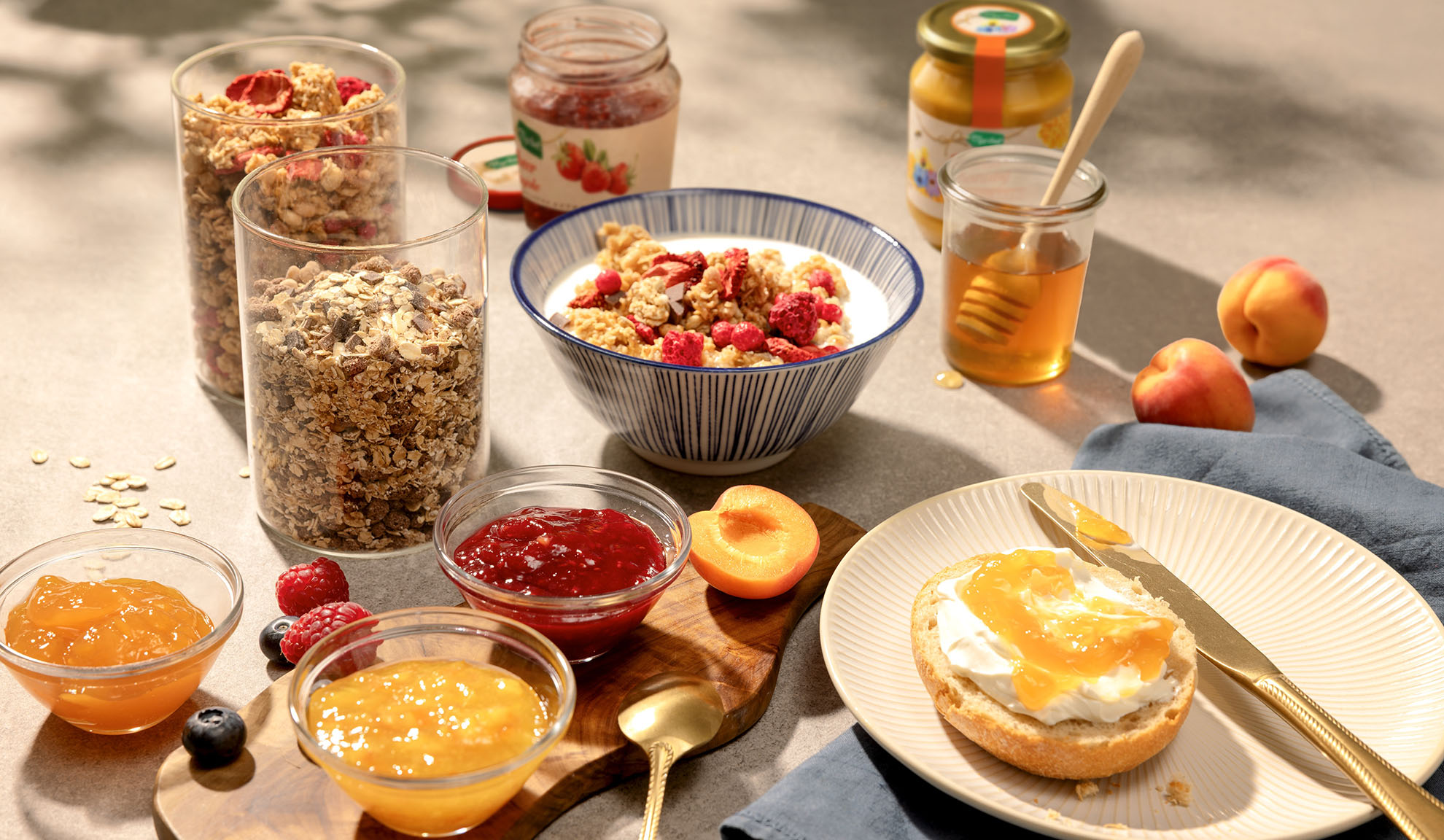 Frühstück mit Müsli, Joghurt, Honig, Marmeladen und frischem Obst auf einem sonnigen Tisch.