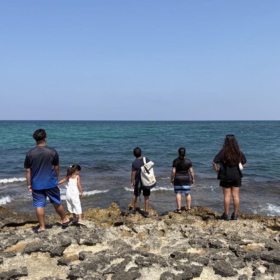 Fünf Personen, darunter ein Kind, stehen am felsigen Ufer und blicken auf das Meer.