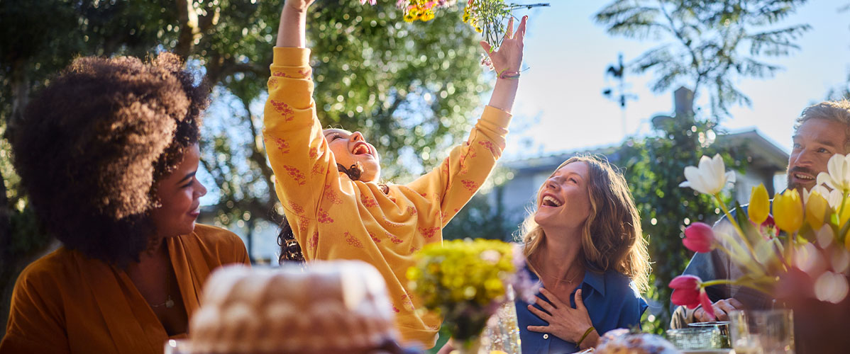 Familie sitzt beim Osterbrunch, die Sonne scheint und Kind wirft glücklich Blumen in die Luft