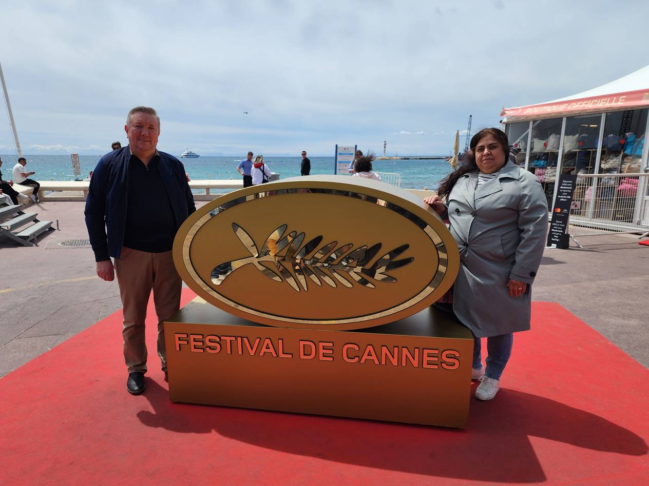 Zwei Personen posieren vor einem großen goldenen Emblem mit der Aufschrift „FESTIVAL DE CANNES“ auf einem roten Teppich.