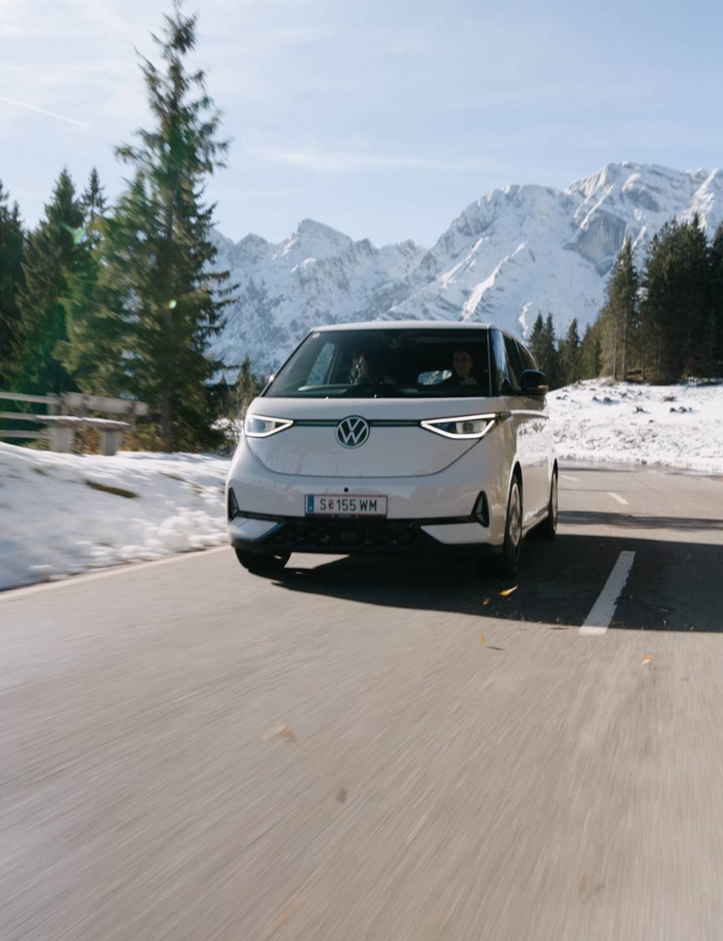 Weißer VW ID. Buzz fährt auf einer Bergstraße mit Schnee und Bäumen im Hintergrund.