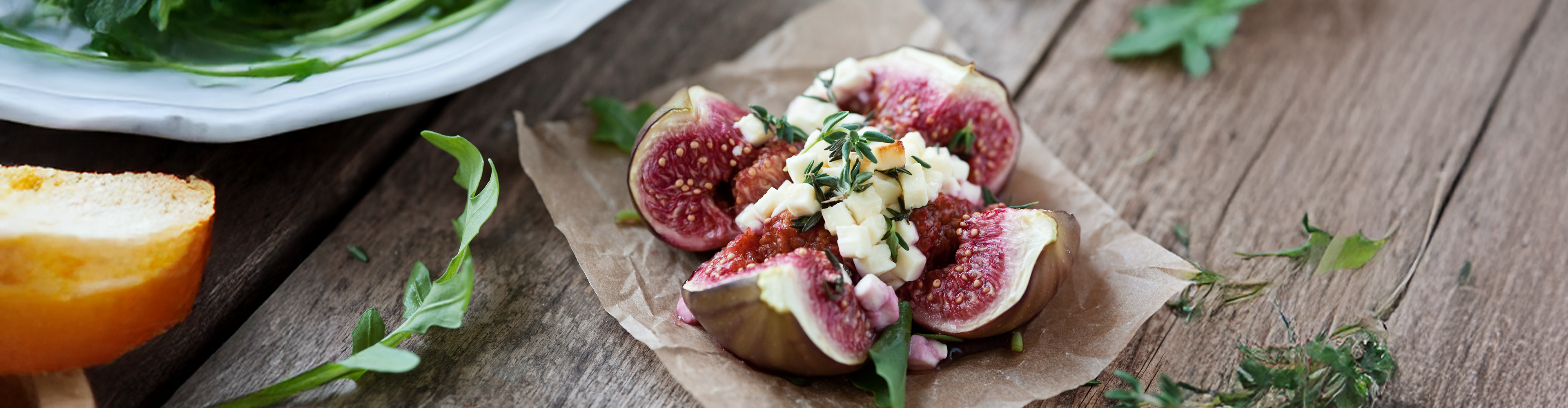 Frische Feigen mit Käse und Kräutern auf Backpapier, daneben Brot und Salat.