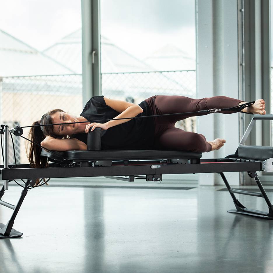 Frau macht Pilates auf einem Reformer, seitlich liegend mit Beingurten.