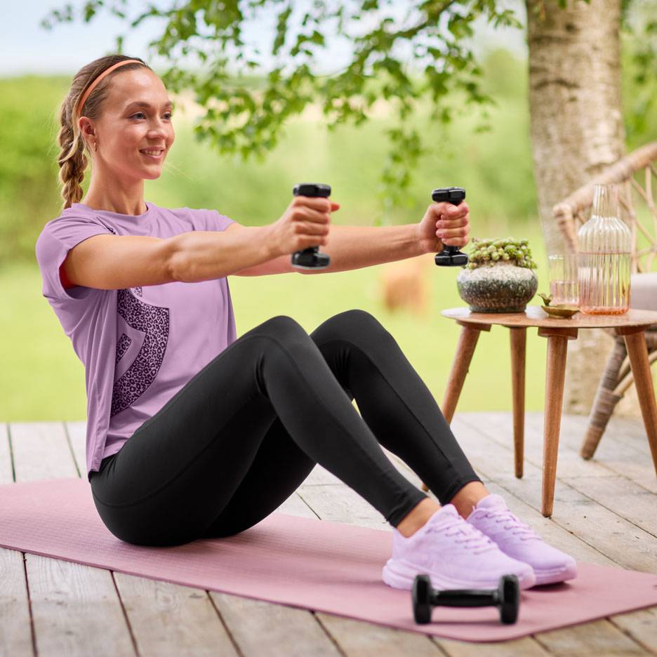 Frau in lila T-Shirt und schwarzen Leggings trainiert mit Hanteln auf einer Yogamatte.