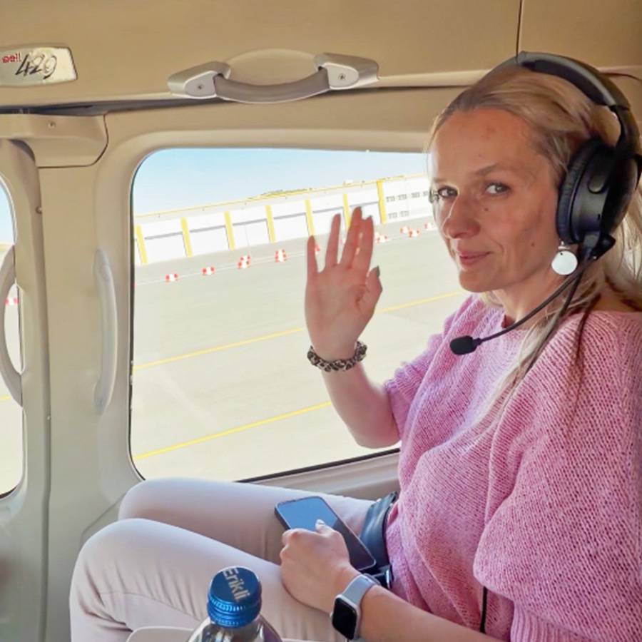 Frau mit Headset winkt aus einem Hubschrauber, hält ein Smartphone und trägt eine Smartwatch.