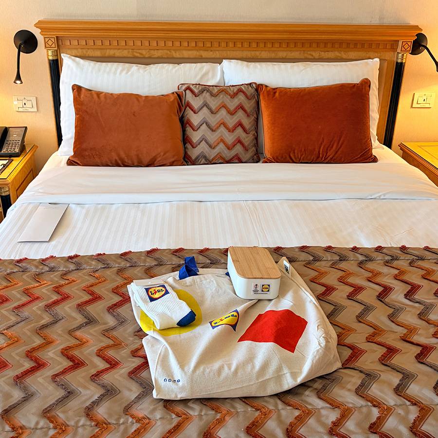 Einkaufstasche, Socken und Brotdose mit Supermarkt-Logo auf einem Hotelbett.