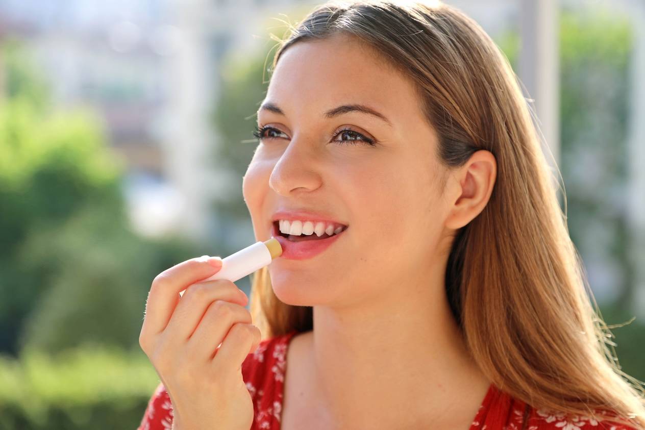 Junge Frau trägt Lippenbalsam auf, lächelnd in der Sonne.