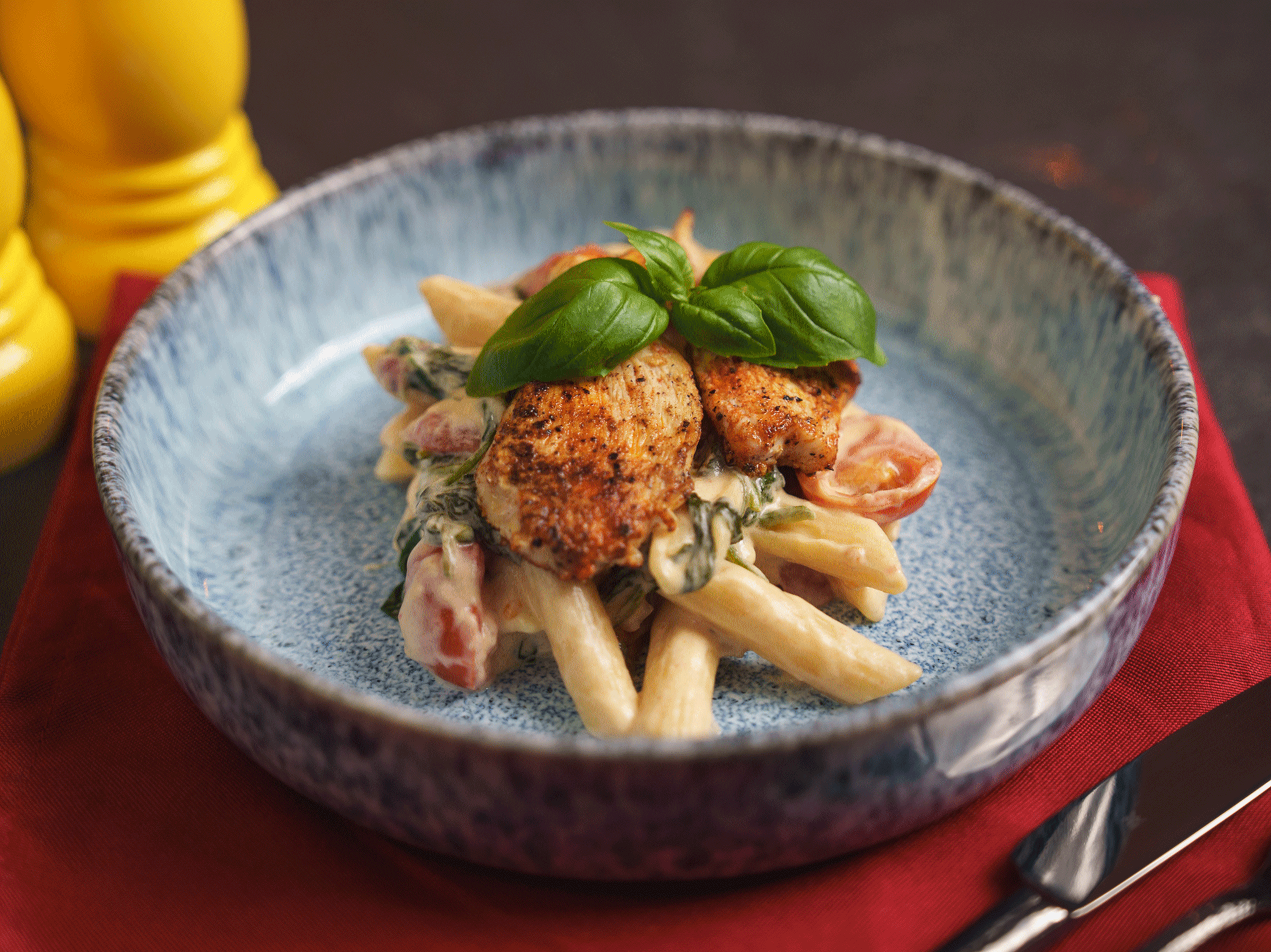 Hähnchen-Penne-Pasta mit Tomaten, Spinat und Basilikum in einer blauen Schale auf rotem Tuch.