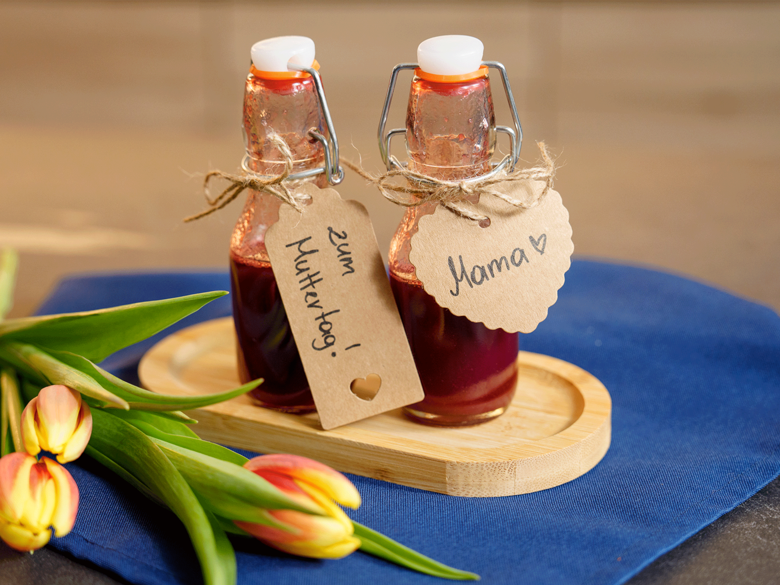 Zwei Flaschen mit rotem Likör und Etiketten 'Zum Muttertag!' und 'Mama ❤️', daneben Tulpen.