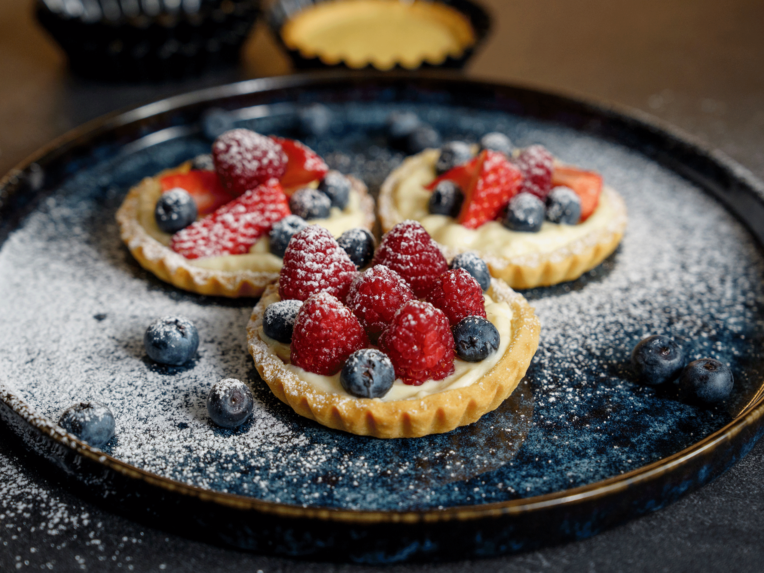 Drei Beeren-Törtchen mit Puderzucker auf einem blauen Teller.