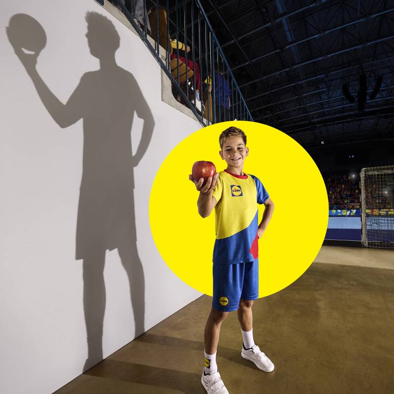 Junge in Lidl Sportkleidung hält Apfel, mit Schatten eines Handballspielers an der Wand.