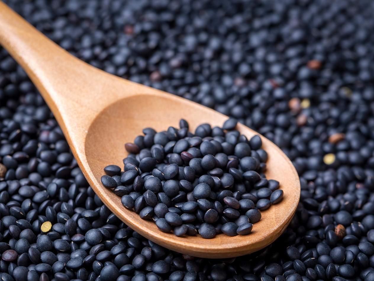 Schwarze Linsen in einem Holzlöffel, umgeben von weiteren Linsen.