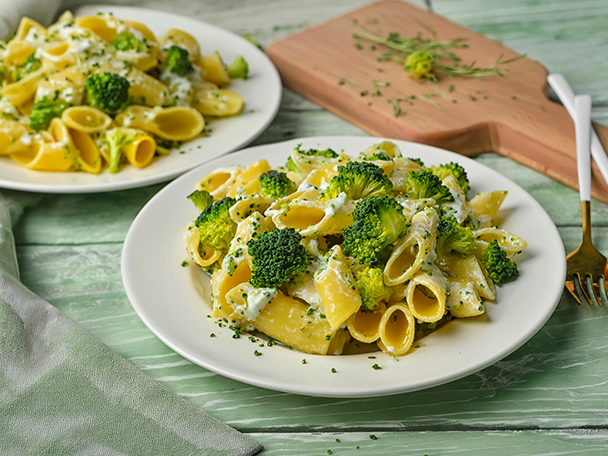 Zwei Teller mit Penne-Nudeln, Brokkoli und cremiger Soße auf einem grünen Holztisch.