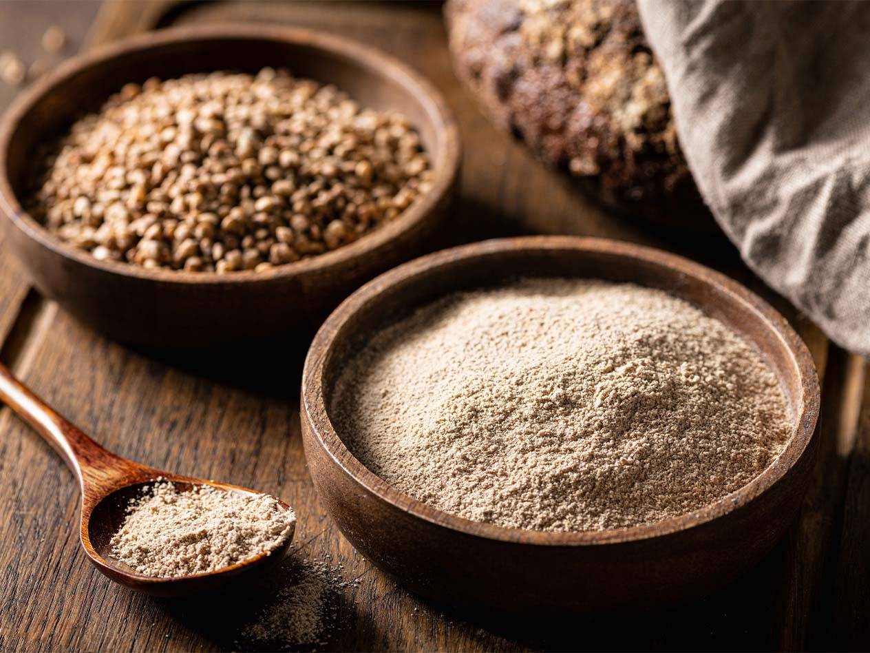 Zwei Holzschalen mit Buchweizen und Buchweizenmehl, daneben ein Laib Brot.