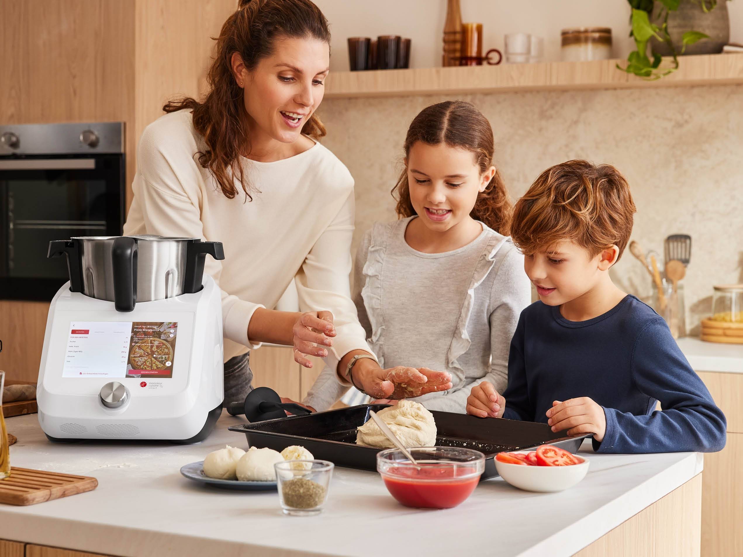 Eine Frau und zwei Kinder bereiten in einer modernen Küche Pizza mit einer Küchenmaschine zu.
