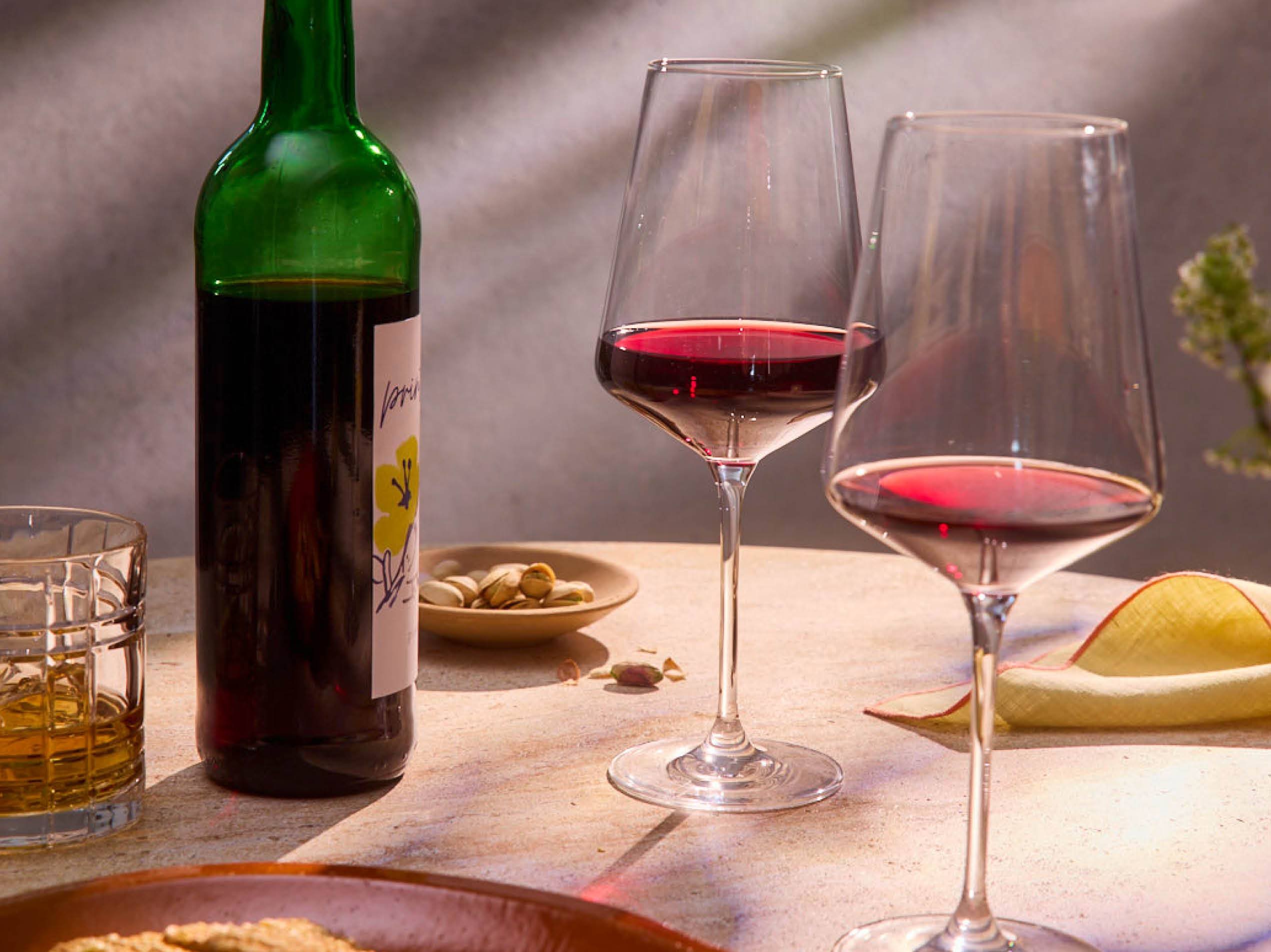 Zwei Gläser Rotwein stehen neben einer Flasche Wein und einer Schale Pistazien auf einem Tisch.