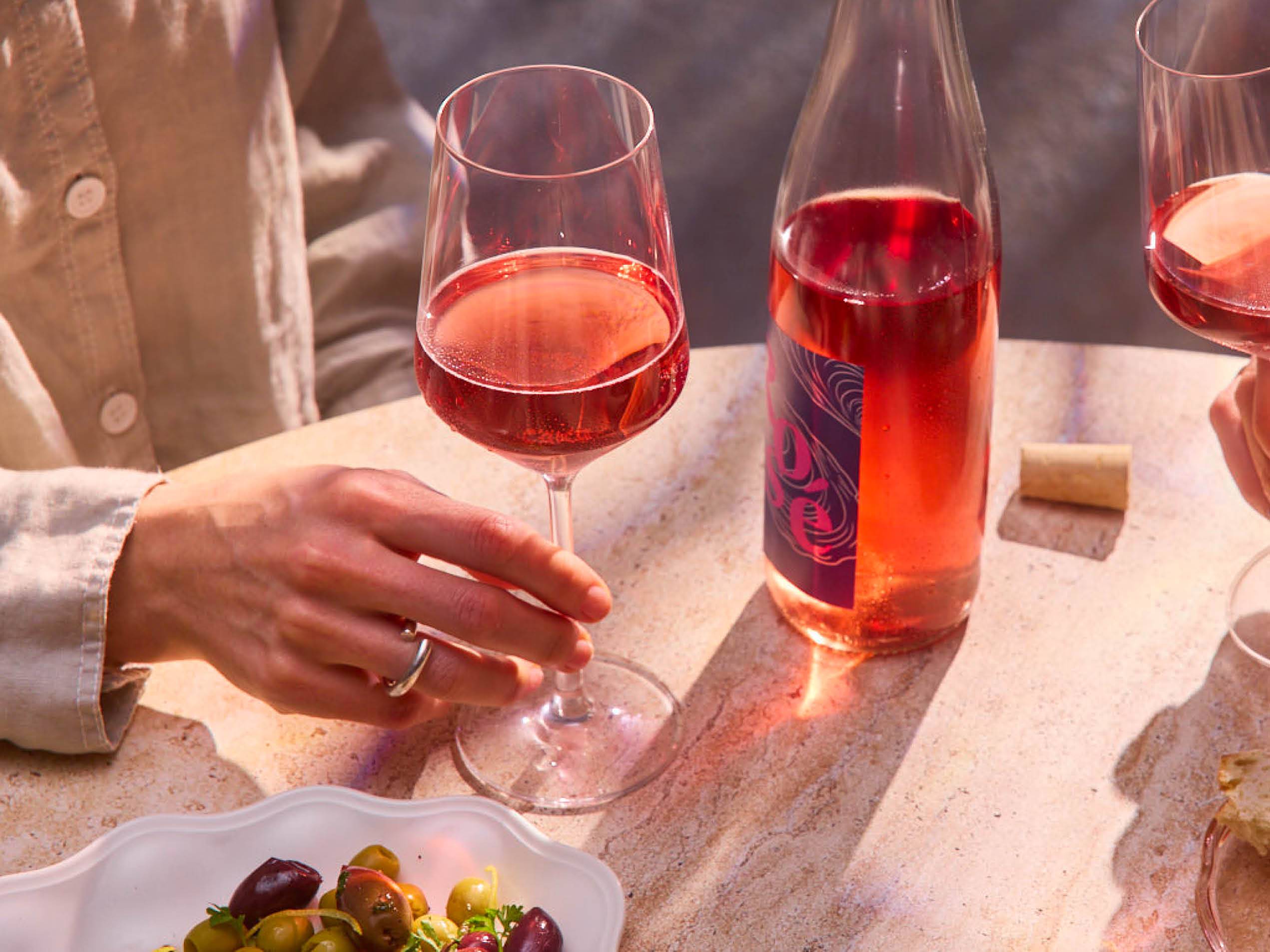 Eine Person hält ein Glas Roséwein daneben steht eine Flasche Wein am Tisch.