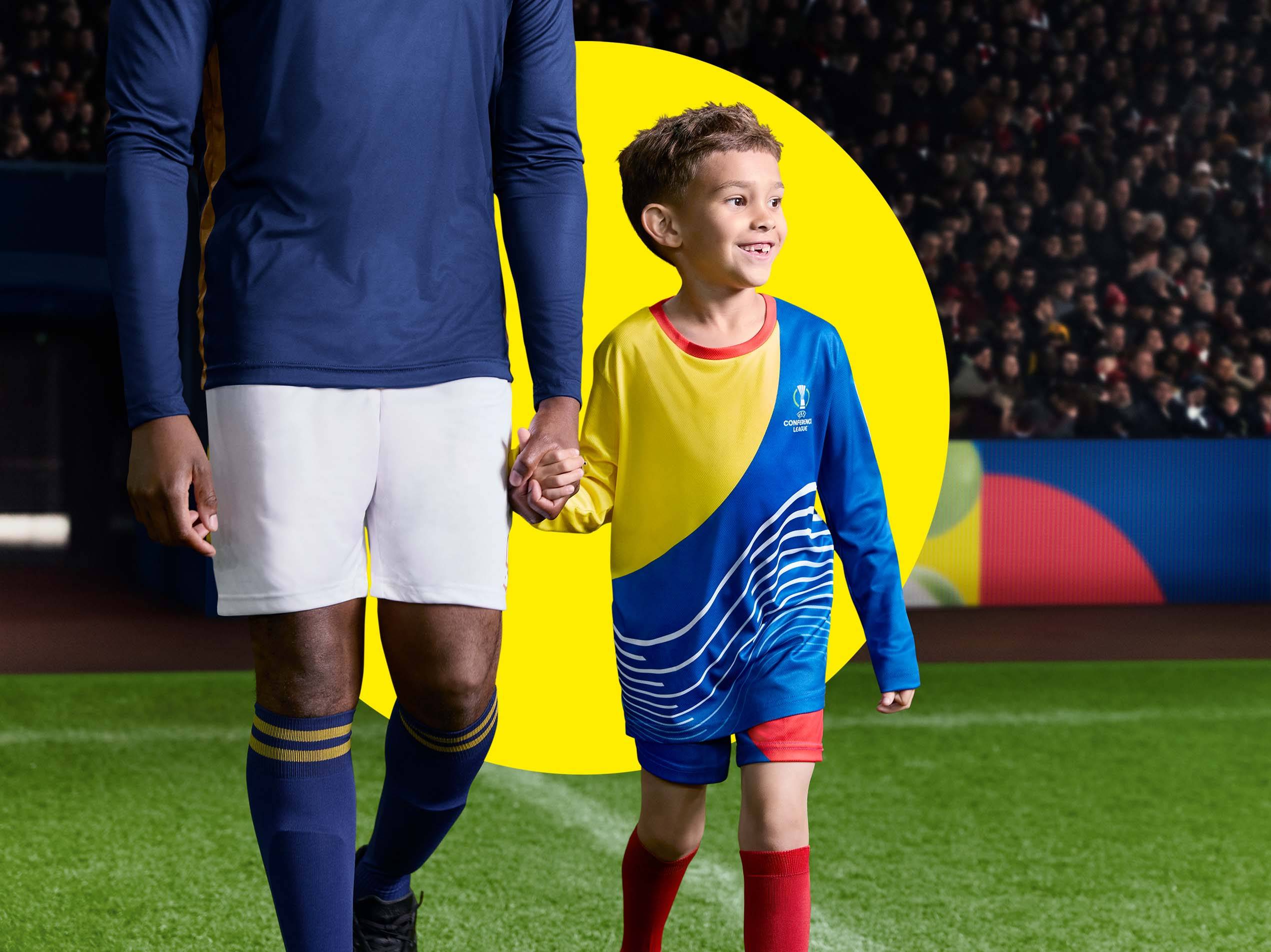 Ein Fußballspieler und ein Kind in Trikots halten Händchen auf einem Fußballfeld.