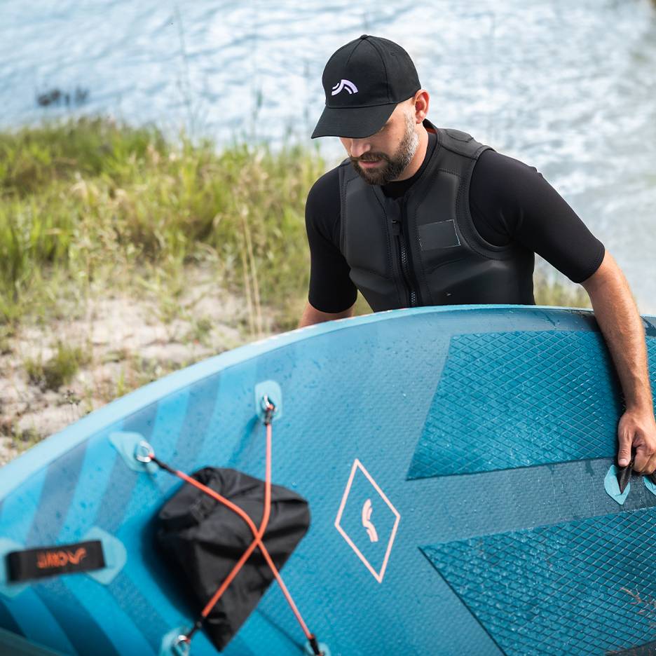 Mann in Schwimmweste und Kappe hält blaues Paddleboard am Wasser.