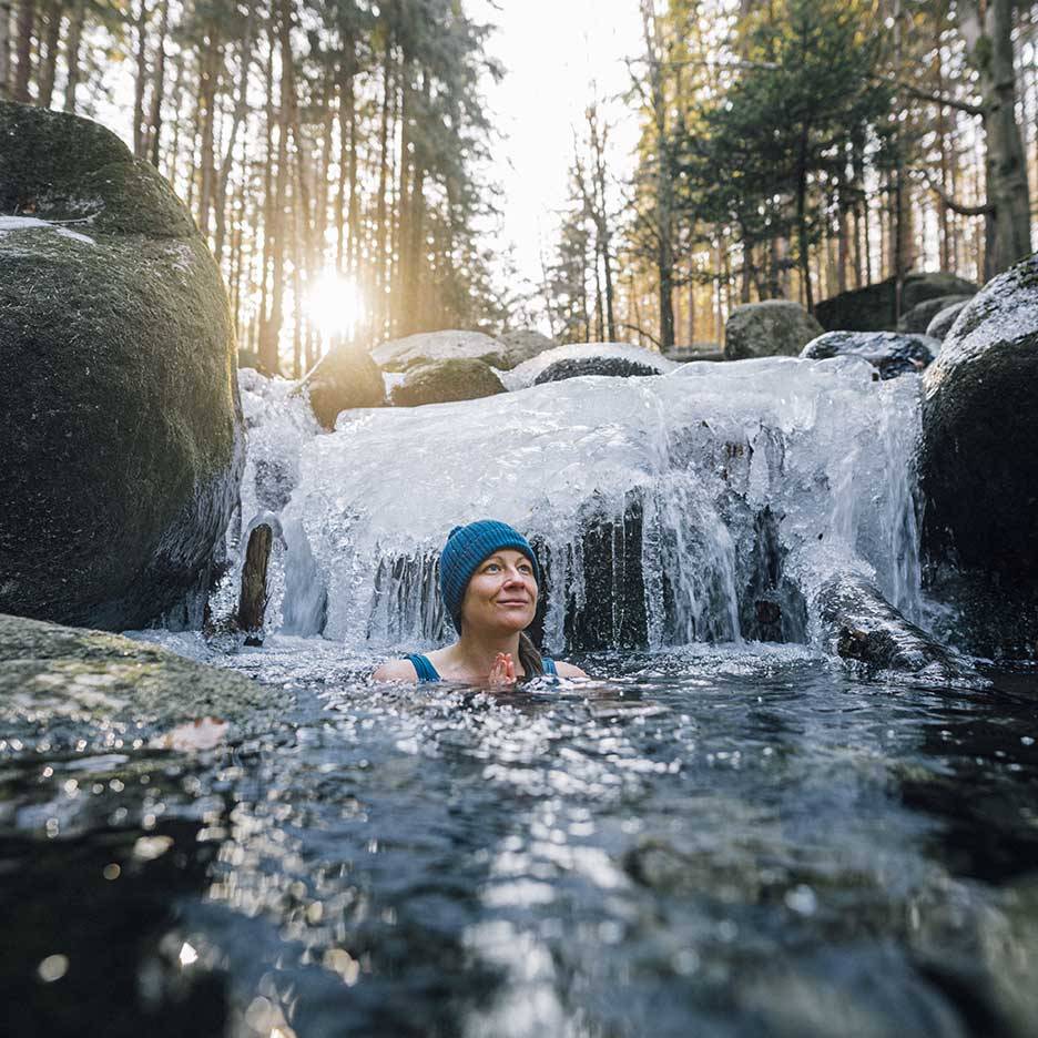 Frau in blauer Mütze und Badeanzug genießt ein kaltes Bad in einem gefrorenen Fluss.