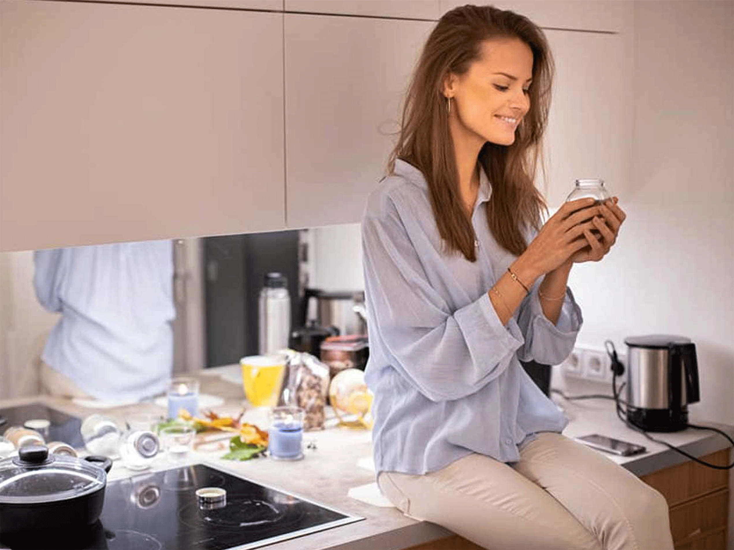 Lächelnde Frau sitzt auf einer Küchenarbeitsplatte und hält ein Glasgefäß.