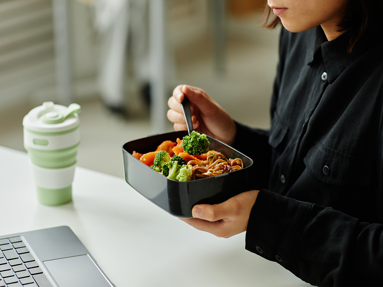 Person isst Nudeln mit Brokkoli aus einer Lunchbox, daneben ein faltbarer Becher.