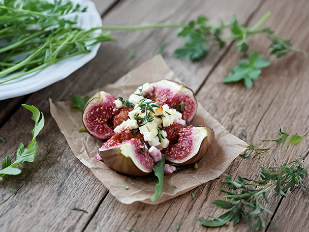 Aufgeschnittene Feigen mit Käsewürfeln und Kräutern auf Backpapier, daneben Rucola.