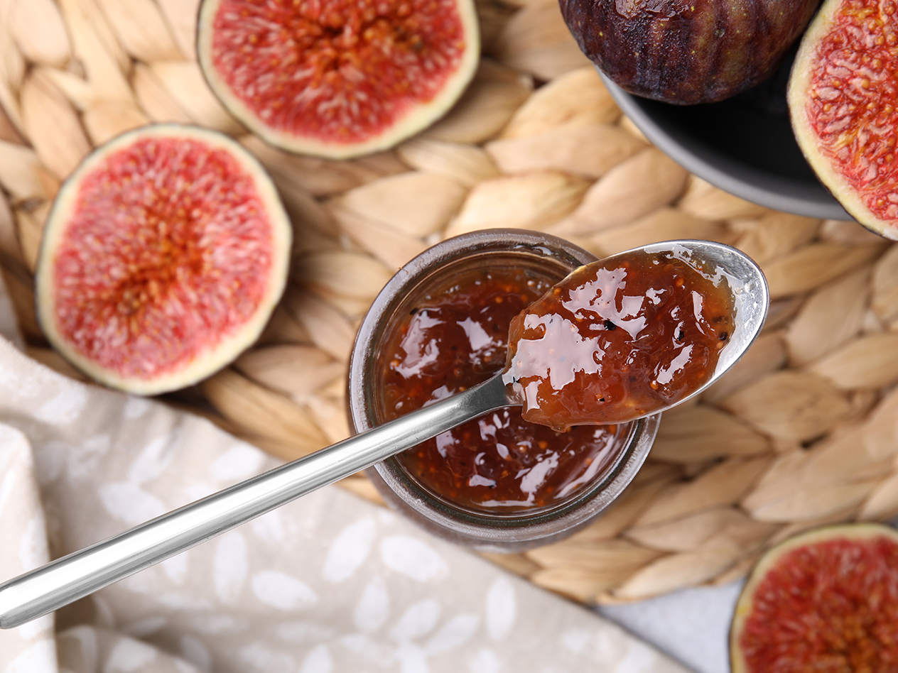 Feigenmarmelade in einem Glas mit einem Löffel, umgeben von frischen Feigen auf einem geflochtenen Untergrund.