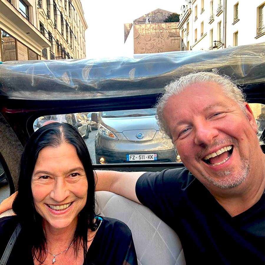 Ein Paar lacht in einem Auto, mit einem anderen Auto im Hintergrund auf einer Straße.